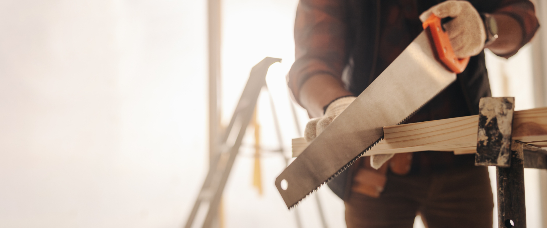 Person cutting a wooden plank with a handsaw on a workbench, wearing gloves and flannel shirt; a stepladder is in the background.